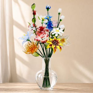 以下が含まれることがあります： 透明なガラスの花瓶に、赤、黄、青、オレンジ、白など、さまざまな色の手作りのガラスの花束が入っています。このアレンジメントは、緑色のガラスの茎と葉を持つ多様な花の形を特徴としています。