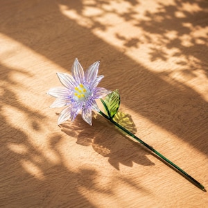 以下が含まれることがあります： 半透明の薄紫色の花びら、小さな黄色い点が散りばめられた淡い青色の中心部、そして側面に一枚の緑色の葉がついた細い緑色の茎を持つ、手作りのガラス製の花のオーナメントです。繊細な花の彫刻のようなデザインです。
