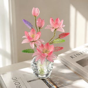 以下が含まれることがあります： 薄いピンク色の半透明な蓮の花と緑の葉が細い茎についた装飾的なアレンジメントで、透明な多面体のクリスタル花瓶に生けられています。花はつぼみから完全に開いた花びらまで、さまざまな開花段階を見せています。