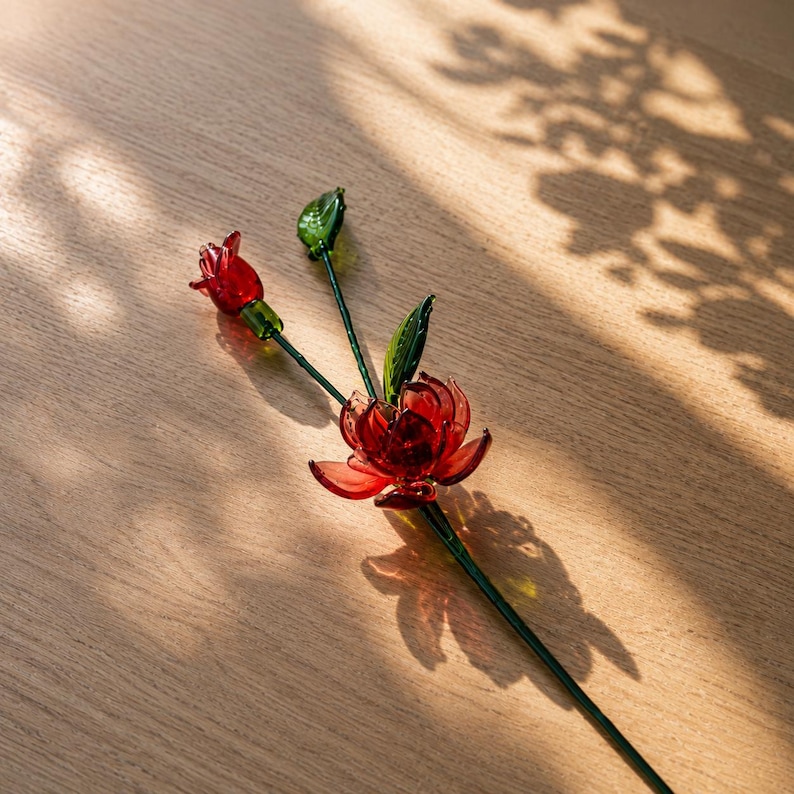 以下が含まれることがあります： 赤い花びらを重ねた大きな花と、小さな赤い蕾、そして2枚の緑の葉が細いダークグリーンのガラスの茎に付いたガラス製の花の彫刻。半透明の赤と緑のガラスで作られており、茎と葉、花びらの質感が表現されています。