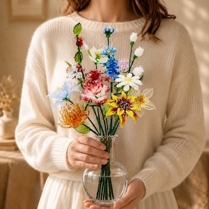 May include: A clear glass vase containing a bouquet of multi-coloured glass flowers. The arrangement features various blooms in shades of red, yellow, blue, and white, each with green glass stems and leaves, creating a vibrant and detailed floral display.