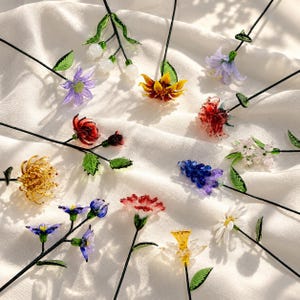 以下が含まれることがあります： 細い黒い茎に手作りされたガラスの花のコレクション。黄色いヒマワリ、赤いカーネーション、青いヒヤシンス、白いデイジー、紫のユリなど、繊細な緑のガラスの葉が付いた様々な種類の花が並んでいます。