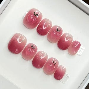 Könnte beinhalten: Ein Set handgefertigter Press-On-Nägel in einer weißen Box. Die Nägel haben ein Farbverlaufsdesign von hellrosa bis zu einer tieferen Rosafarbe, mit kleinen schwarzen floralen Akzenten. Die Box trägt den Text "HANDMADE NAIL" und die Nummer "19740".