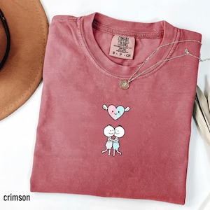 May include: Crimson-colored t-shirt with a crew neck, featuring a heart with wings and two hugging figures. The shirt has a Comfort Colors tag and a silver necklace with a pendant. The word "crimson" is at the bottom left.