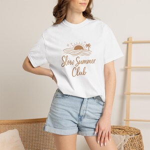 May include: White t-shirt with a brown graphic of a sun, waves, and a palm tree, with the text "Slow Summer Club". The shirt is paired with denim shorts. The overall theme is casual summer wear.