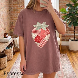 May include: A maroon-coloured t-shirt with a large patchwork strawberry design. The strawberry is made from various red and white patterned fabrics, with a green leaf detail. The word "Espresso" is at the bottom. The shirt is a loose fit.