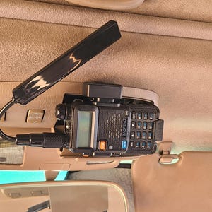 May include: A black and gray Baofeng radio transceiver mounted inside a vehicle. The radio has a small screen, a keypad, and various buttons. A black antenna is attached to the radio. The radio is mounted on a beige surface.