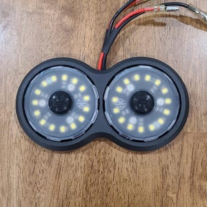 May include: A dual-lens automotive light assembly featuring two circular LED arrays housed in a black matte frame. Each lens contains a ring of small white LED bulbs and a central black circular element. Red and black wires extend from the top.
