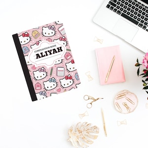 Puede incluir: Un cuaderno de composición rosa con un diseño de Hello Kitty y el nombre "ALIYAH" en un marco blanco. La imagen incluye un portátil, un cuaderno rosa y accesorios dorados sobre una superficie blanca.