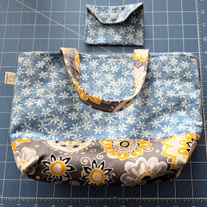 以下が含まれることがあります： 手作りのトートバッグと、お揃いの小さな財布。バッグは、白の花柄とグレーと黄色の花のアクセントが入った水色です。財布はボタン留めで、バッグの花柄とマッチしています。バッグには「HANDMADE」と書かれた小さなタグが付いています。