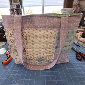 Puede incluir: Un bolso tote acolchado grande con un diseño de patchwork. El bolso presenta varios paneles de tela floral y de lunares en tonos lavanda, crema y azul. Las asas son de color lavanda liso.