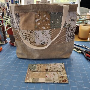 Könnte beinhalten: Eine Patchwork-Tragetasche mit passendem Portemonnaie. Die Tasche hat eine beige Canvas-Basis und ein Patchwork-Design mit floralen und gepunkteten Mustern in Beige, Braun und Grün. Das Portemonnaie hat ein ähnliches Patchwork-Design. Beide Artikel befinden sich auf einer blauen Oberfläche.
