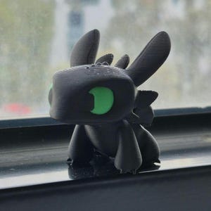 Peut inclure: Figurine de dragon noir imprimée en 3D avec de grands yeux verts. Le dragon a un design stylisé avec des oreilles proéminentes et un petit corps. La figurine est posée sur un rebord de fenêtre.