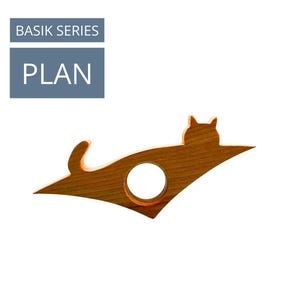 May include: A wooden cat-shaped book holder with a circular opening. The cat design is in a lying down position. The text "BASIK SERIES" and "PLAN" are in a blue rectangle in the upper left corner. The book holder is made of light brown wood.