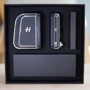 Könnte beinhalten: Eine schwarze Box mit einem schwarzen, ledertexturierten Artikel mit silbernem Rand und dem Buchstaben "H", einem kleineren schwarzen Artikel und einem schwarzen rechteckigen Artikel. Die Artikel sind in einer schwarzen Box angeordnet.