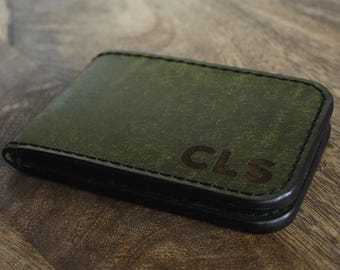 May include: Green leather wallet with black stitching and the initials "CLS" embossed on the front. The wallet is rectangular and appears to be made of high-quality leather. The background is a wooden surface.