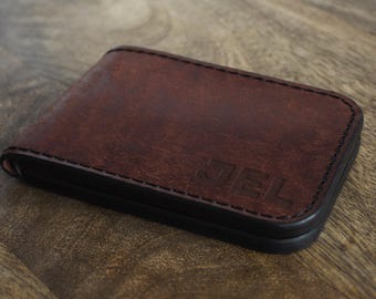 May include: A brown leather wallet with a black border. The wallet has the initials "JEL" embossed on the front. The wallet is rectangular and appears to be made of high-quality leather.