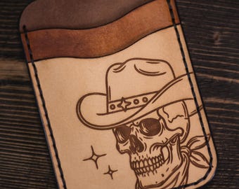 May include: A tan leather cardholder with a cowboy skull design. The cardholder has three slots and a brown top. The skull is wearing a cowboy hat and bandana. The design is etched into the leather.