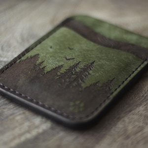 May include: A leather card holder with a forest scene. The design features dark brown trees against a green background, with a dark brown border. Two birds are flying in the sky. The card holder is rectangular and has a stitched edge.