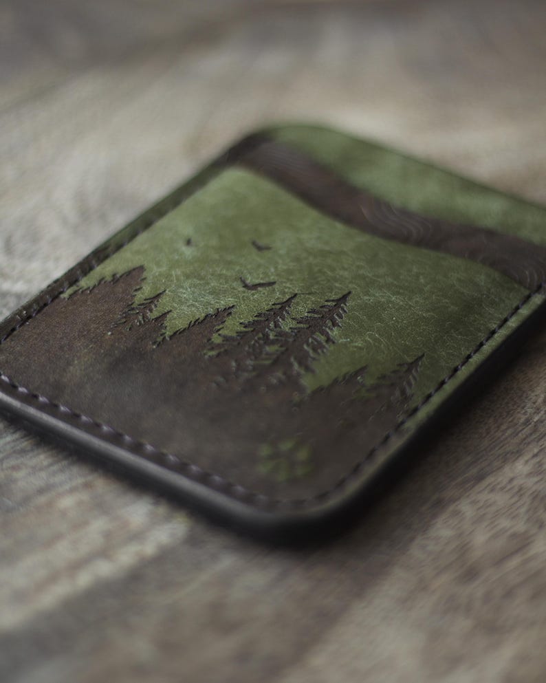 May include: A leather card holder with a forest scene. The design features dark brown trees against a green background, with a dark brown border. Two birds are flying in the sky. The card holder is rectangular and has a stitched edge.
