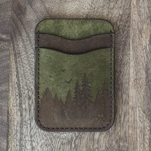 May include: Green leather cardholder with a forest scene design. The cardholder features a dark brown accent and a stitched border. The forest scene includes trees and birds. The cardholder is rectangular and has a paw print symbol.