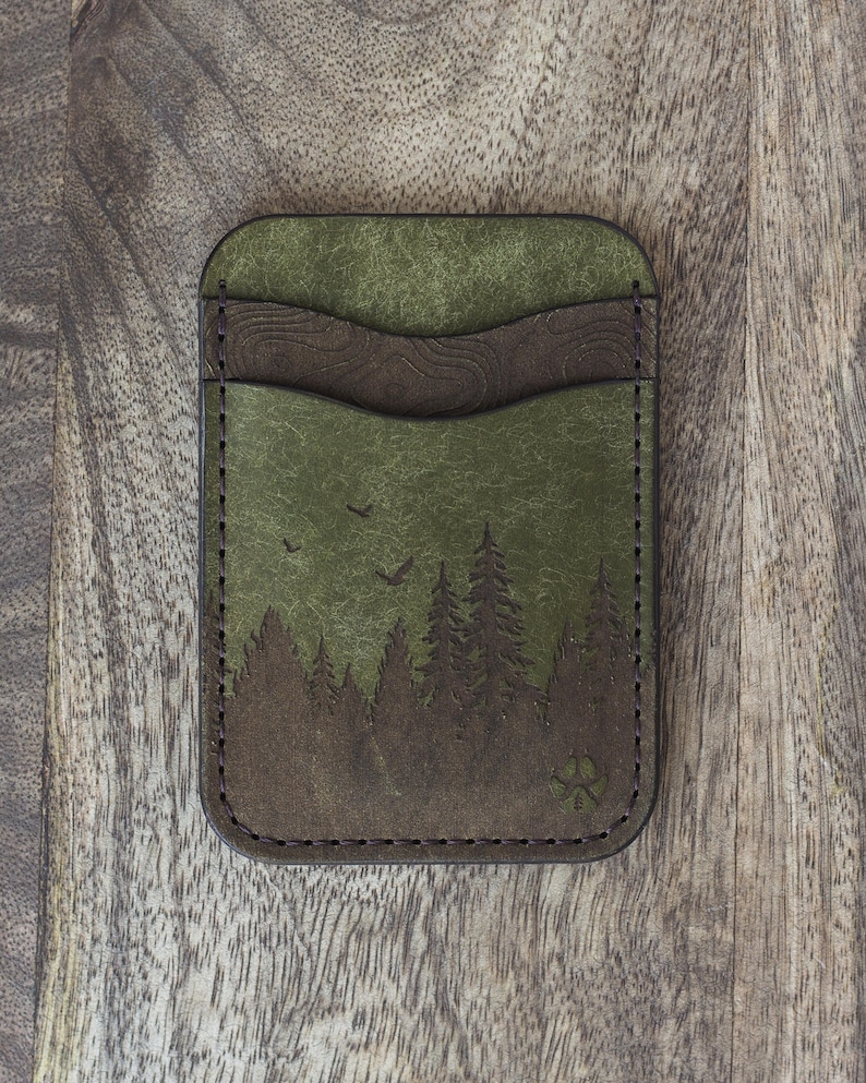 May include: Green leather cardholder with a forest scene design. The cardholder features a dark brown accent and a stitched border. The forest scene includes trees and birds. The cardholder is rectangular and has a paw print symbol.