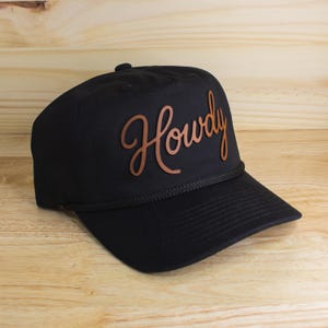 Op de afbeelding: Zwarte baseballcap met een gebogen klep en een touwdetail. Het woord "Howdy" staat in bruin, cursief lettertype op de voorkant. De cap staat tegen een houten achtergrond.