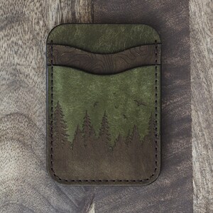May include: Green and brown leather cardholder with a forest scene. The cardholder features a detailed forest silhouette with trees and birds, and a textured brown accent. The edges are stitched with dark thread.