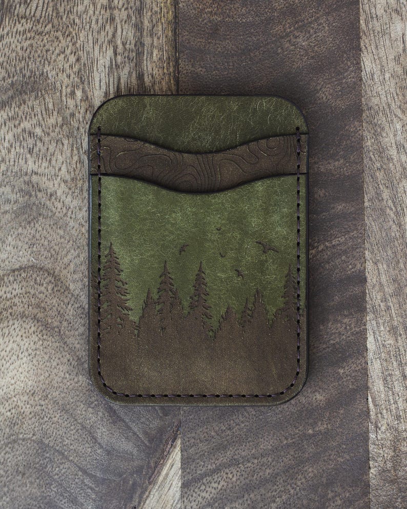 May include: Green and brown leather cardholder with a forest scene. The cardholder features a detailed forest silhouette with trees and birds, and a textured brown accent. The edges are stitched with dark thread.