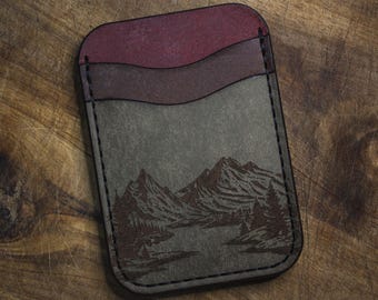 May include: A leather card wallet with a mountain and lake scene etched on the front. The wallet has three pockets in shades of burgundy, brown, and gray. The edges are stitched with black thread.
