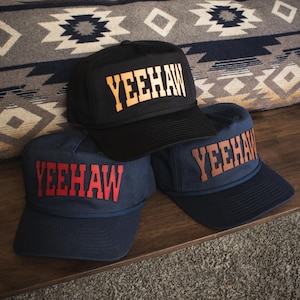 Puede incluir: Tres gorras de béisbol con la palabra "YEEHAW" en letras en negrita. Una gorra es negra con texto naranja, y dos son azul marino con texto rojo y naranja. Las gorras se exhiben sobre una superficie de madera.