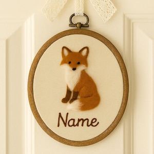 May include: A circular embroidery hoop featuring a felt fox design. The fox is depicted in shades of brown and white, set against a cream fabric. The word "Name" is embroidered below. A cream lace bow is suspended above the hoop.