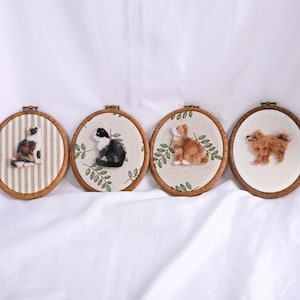 May include: Four oval-shaped, wooden framed embroidery hoops with felted animal designs. The designs include a calico cat on striped fabric, a black and white cat with greenery, a brown rabbit with greenery, and a brown dog.