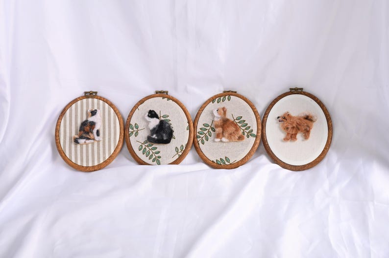 May include: Four oval-shaped, wooden framed embroidery hoops with felted animal designs. The designs include a calico cat on striped fabric, a black and white cat with greenery, a brown rabbit with greenery, and a brown dog.