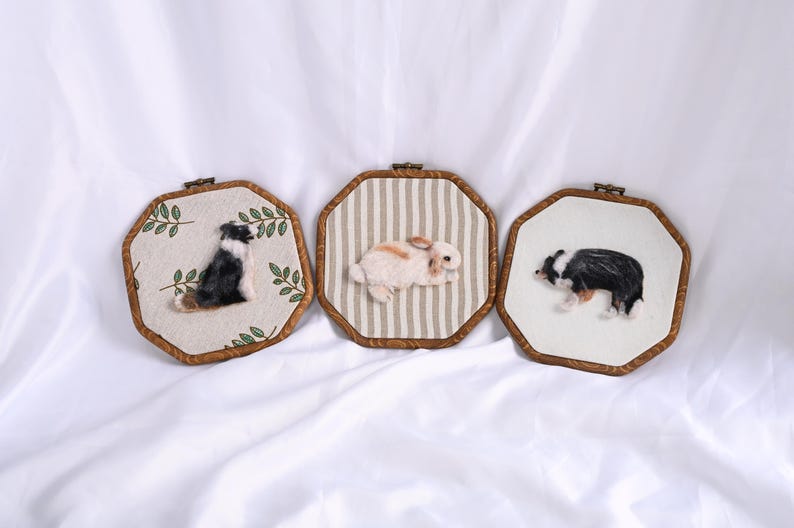 May include: Three framed felted animal portraits. One features a black and white dog with a brown patch, another a brown and white rabbit, and the third a black and white dog. Each is set in a wooden frame.