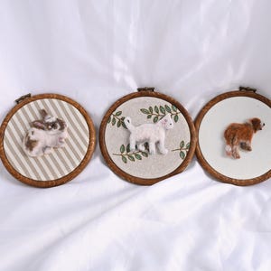 May include: Three framed felt art pieces featuring animals. A rabbit on striped fabric, a white cat with green leaves, and a brown dog on a white background. Each is in a wooden embroidery hoop.