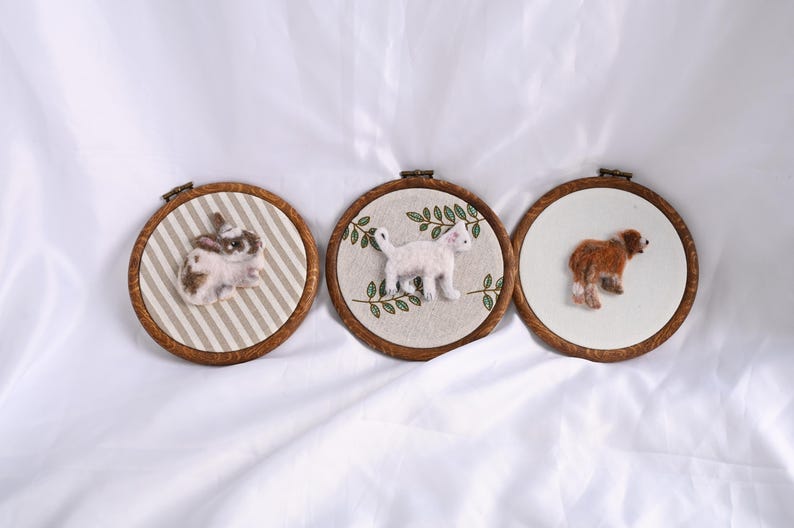 May include: Three framed felt art pieces featuring animals. A rabbit on striped fabric, a white cat with green leaves, and a brown dog on a white background. Each is in a wooden embroidery hoop.