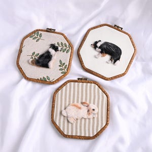 May include: Three handmade wall hangings featuring animal portraits. Two depict black and white dogs, and one shows a brown and white rabbit. Each is framed in a wooden, octagonal hoop with fabric backgrounds.