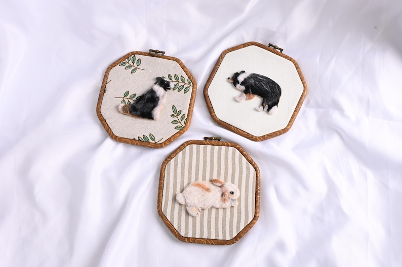 May include: Three handmade wall hangings featuring animal portraits. Two depict black and white dogs, and one shows a brown and white rabbit. Each is framed in a wooden, octagonal hoop with fabric backgrounds.