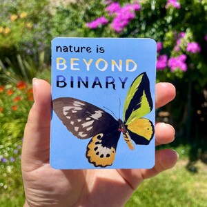 May include: A rectangular sticker with a light blue background featuring a butterfly with black, yellow, and white wings. The text "nature is BEYOND BINARY" is printed in black and white lettering at the top of the sticker, which has rounded corners.