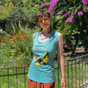 Op de afbeelding: Een mouwloze petrolkleurige tanktop met een afbeelding van een geel-zwarte vlinder en de witte tekst "nature is BEYOND BINARY" op de voorkant. De top wordt gecombineerd met een textuurrijke, roestkleurige rok en een zonnebril met amberkleurige glazen.