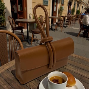 May include: A tan brown rectangular handbag featuring a smooth finish and a distinctive knotted top handle design. The bag has a structured flap closure and a clean, minimalist silhouette, positioned on a wooden table next to a white coffee cup.