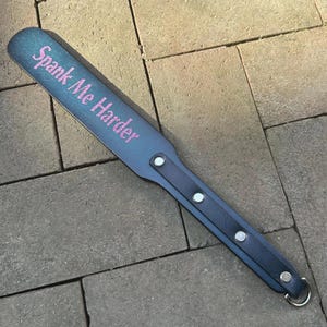Puede incluir: Una paleta azul con el texto "Spank Me Harder" en rosa. La paleta tiene un mango con tres remaches plateados y un anillo de metal en el extremo.