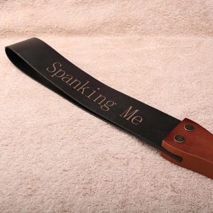 May include: A black leather strap featuring the text Spanking Me engraved in a serif font. The strap is attached to a smooth brown wooden handle by two metallic rivets, creating a long, flexible paddle-like object with a curved design.