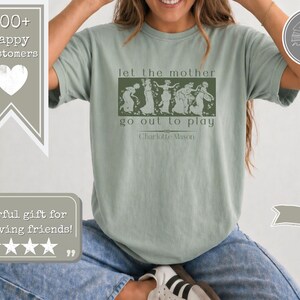 May include: A sage green short-sleeve t-shirt featuring a dark green rectangular graphic with a line of dancing figures. The text reads let the mother go out to play and Charlotte Mason. The shirt is paired with blue denim jeans and white trainers.