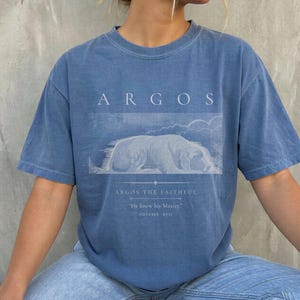 May include: A blue t-shirt with the word "ARGOS" printed at the top. Below the word is a white illustration of a dog lying down, with the text "ARGOS THE FAITHFUL" and a quote from the Odyssey.