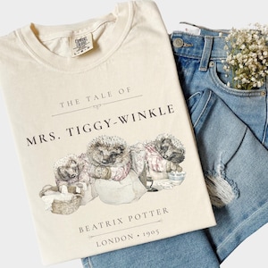 May include: A cream-colored t-shirt featuring a graphic illustration of hedgehogs from The Tale of Mrs. Tiggy-Winkle by Beatrix Potter, with the text "The Tale of Mrs. Tiggy-Winkle" and "Beatrix Potter London 1905" printed in black ink.