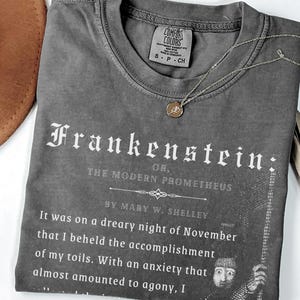 May include: A gray t-shirt with the title "Frankenstein" printed in white gothic font. The text "OR, THE MODERN PROMETHEUS" and a quote from Mary W. Shelley are also printed on the shirt. A gold necklace and a brown hat are visible.