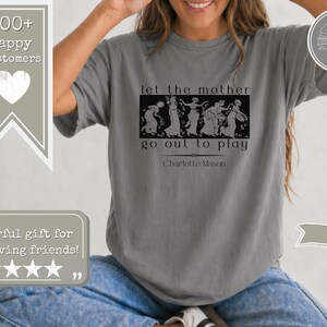 May include: A grey short-sleeved t-shirt featuring a black graphic print of classical figures in a rectangular frame. The text reads let the mother go out to play and Charlotte Mason. The shirt has a crew neckline and a relaxed, loose fit.