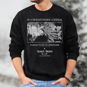 May include: Black sweatshirt featuring a vintage illustration from "A Christmas Carol" by Charles Dickens. The design includes the title, author, and a scene with figures in a graveyard. The text is in white, and the sweatshirt is a solid black color.
